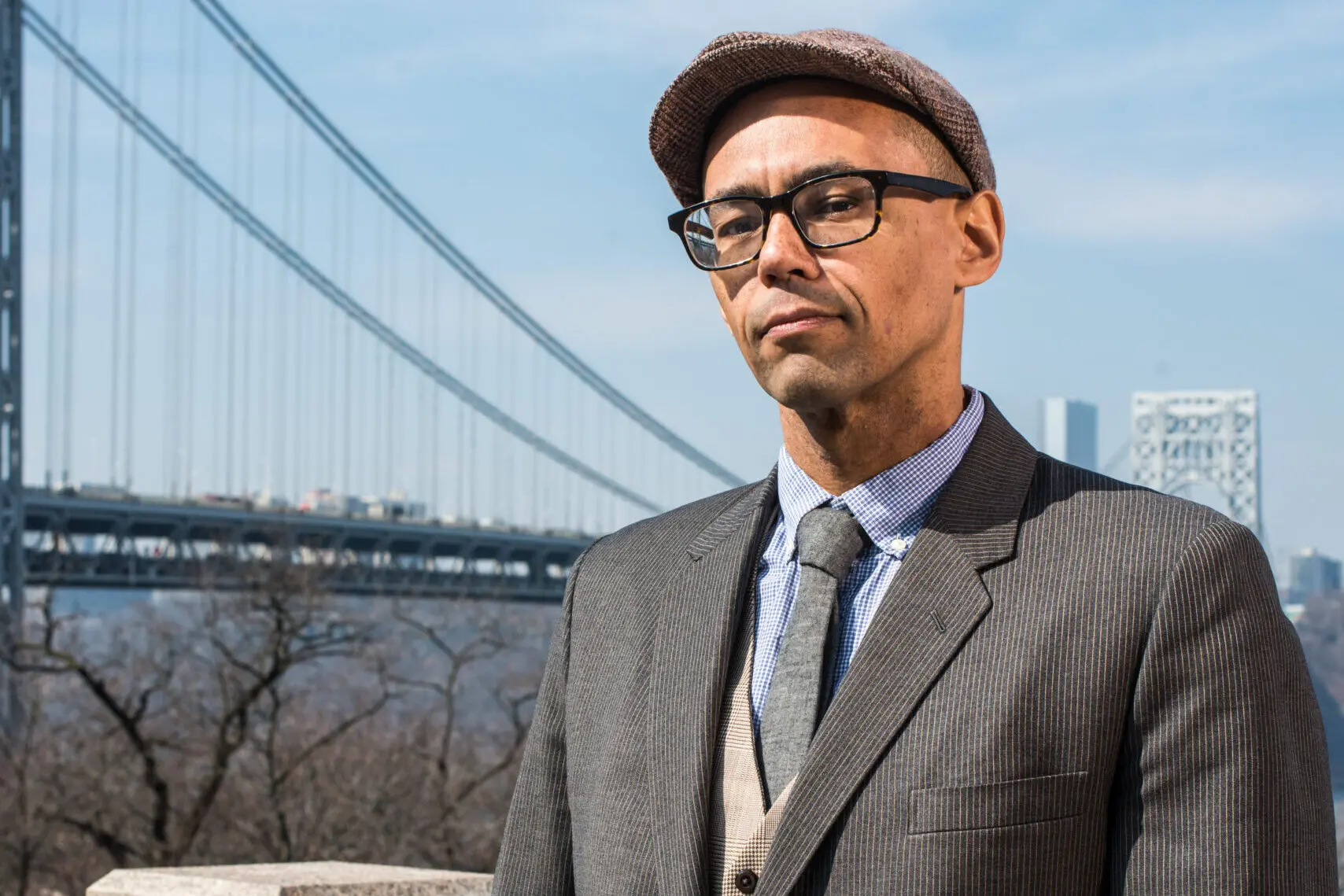 A man wearing glasses, a flat cap, and a suit stands outdoors with a large suspension bridge and city buildings visible in the background on a clear day.