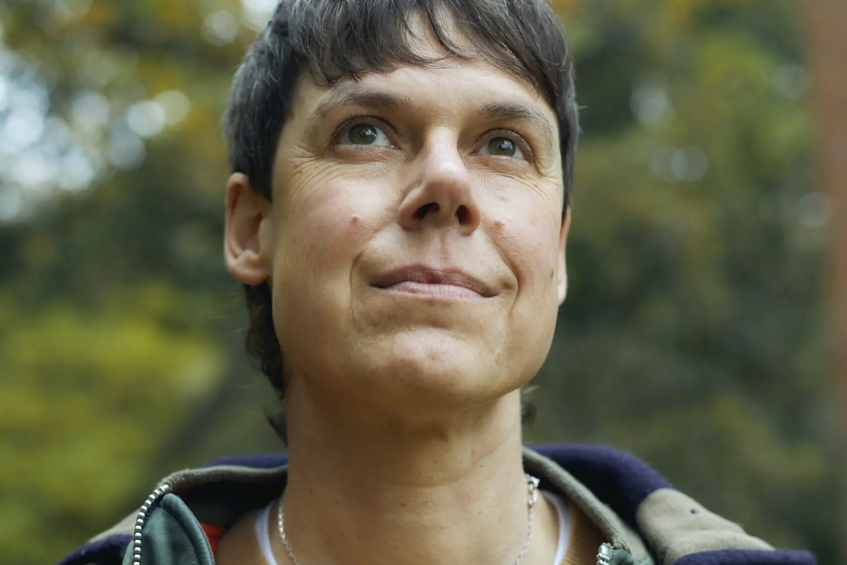 A person with short dark hair and light skin looks upward with a soft smile. They are wearing a jacket and necklace, with a blurred, green outdoor background behind them.