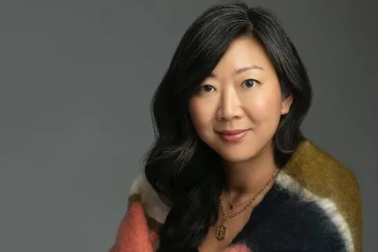 A woman with long, wavy black hair smiles softly at the camera. She is wearing a gold necklace and a colorful, fuzzy shawl draped over her shoulders, with a neutral gray background behind her.