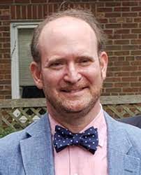 A man with a receding hairline, wearing a light blue blazer, pink shirt, and navy blue polka dot bow tie, smiles in front of a brick wall and window.