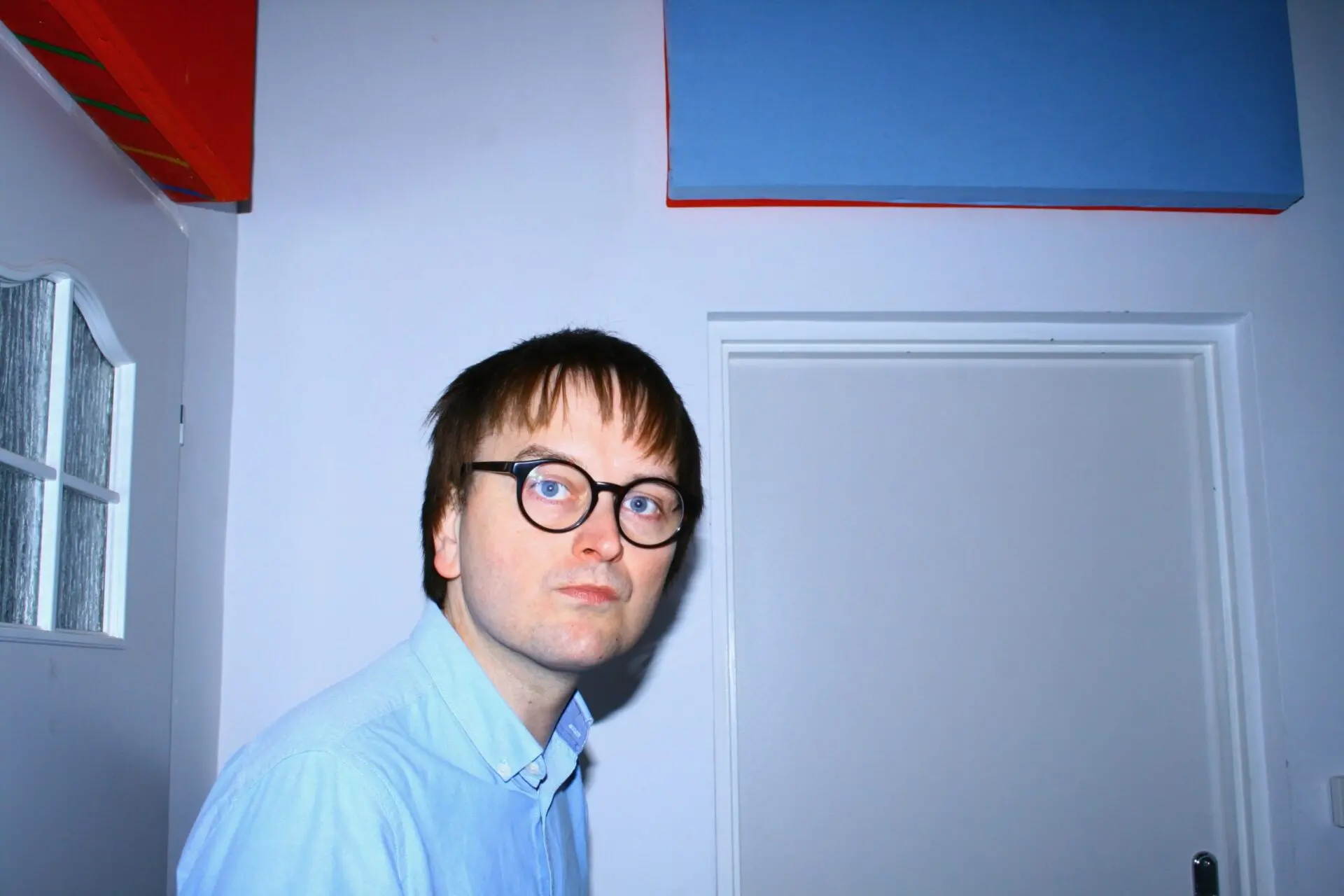 A person with short brown hair and glasses, wearing a light blue shirt, stands indoors near a white door and blue and red panels on the walls. The lighting is bright and the background is plain.