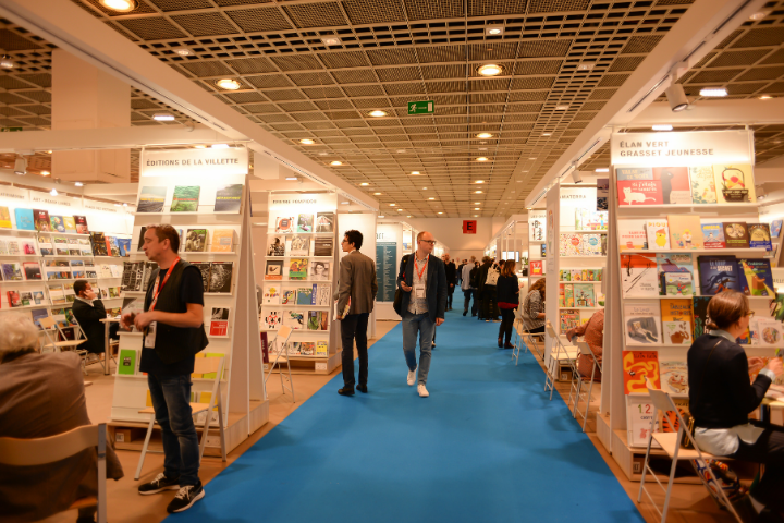 People walk along a blue carpeted aisle at a bustling book fair, with books displayed on shelves from various publishers on both sides. Some visitors sit and browse, while others converse or explore exhibits.