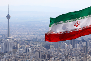 A large Iranian flag waves in the foreground above a cityscape of Tehran, with the Milad Tower visible among many buildings and mountains in the distance.