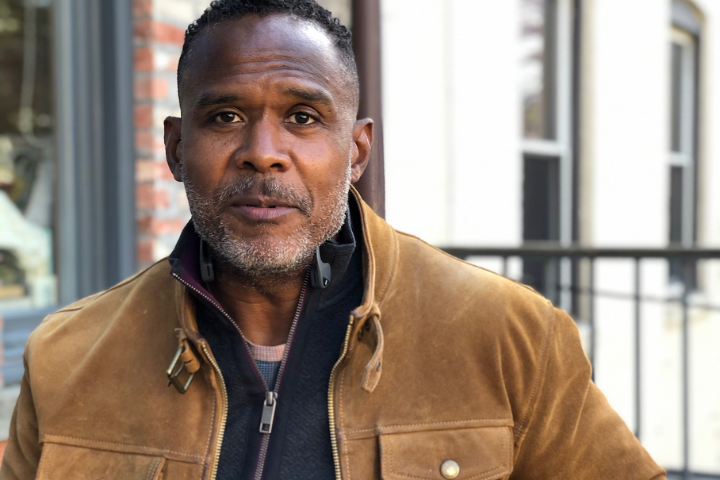 A middle-aged man with short curly hair and a trimmed beard wears a brown suede jacket over a zip-up sweater, standing outdoors in front of a brick wall and windows.