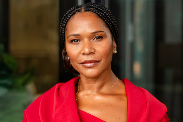 A woman with braided hair, wearing a red, off-the-shoulder top, looks calmly at the camera. She stands in front of a blurred indoor background.