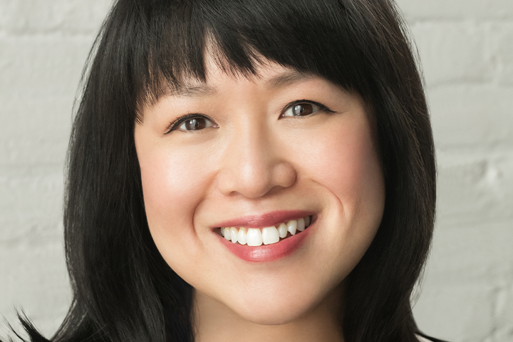 A woman with straight black hair and bangs smiles at the camera. She has clear skin, brown eyes, and is standing in front of a white brick wall.
