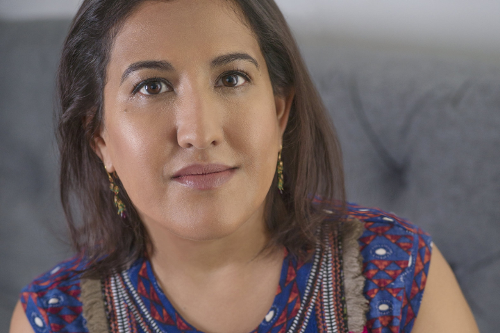 A woman with shoulder-length brown hair wearing a blue patterned top and colorful earrings sits on a gray couch, looking at the camera with a neutral expression.