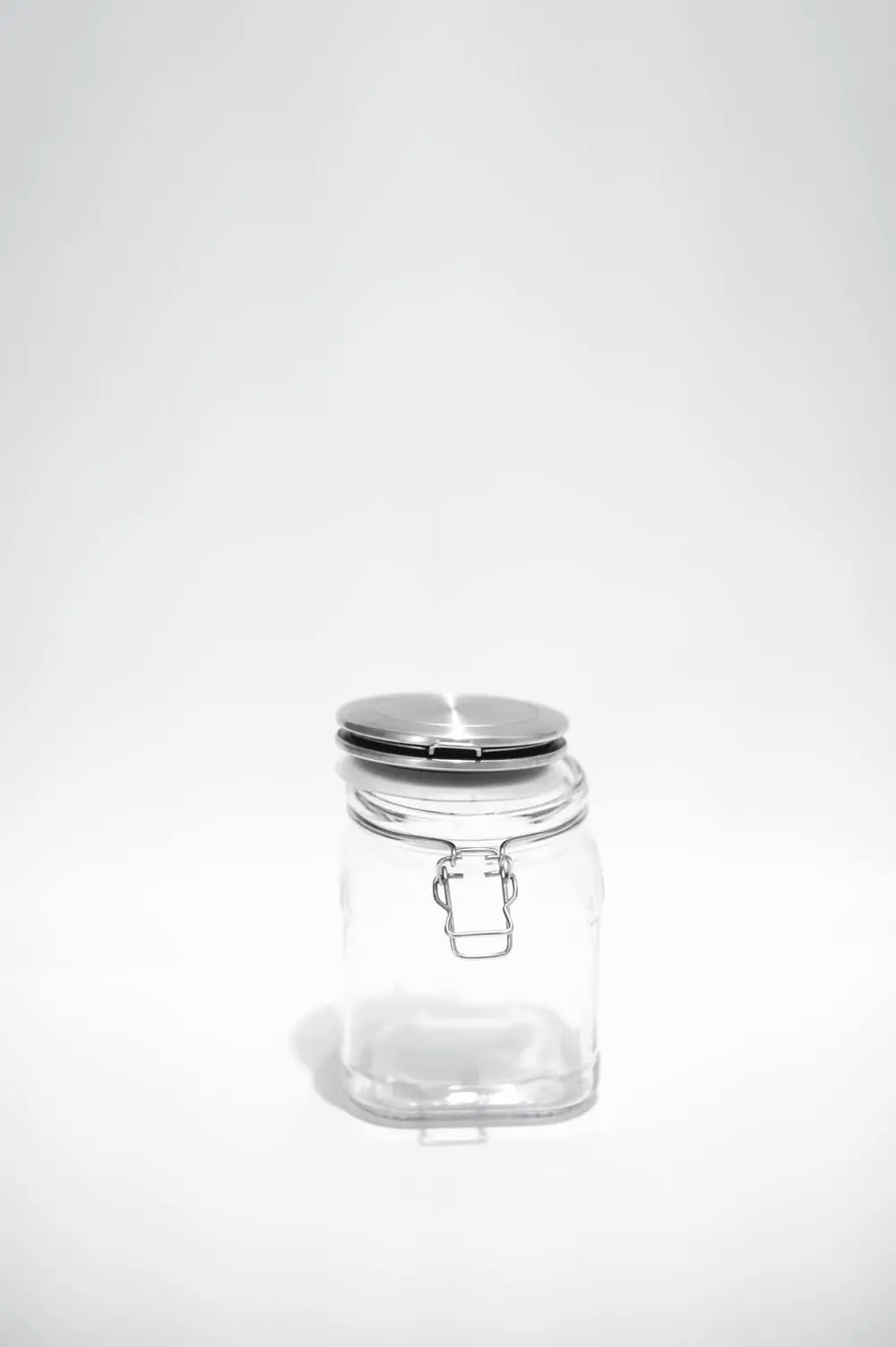 A clear glass jar with a metal clasp and a shiny, silver lid sits on a plain white background. The jar is empty and centered in the image.