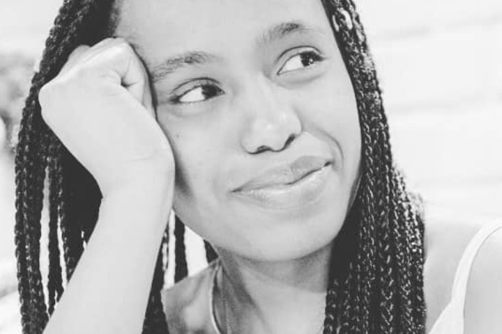 A person with long braids rests their head on their hand and gazes off to the side, smiling softly. The photo is in black and white.