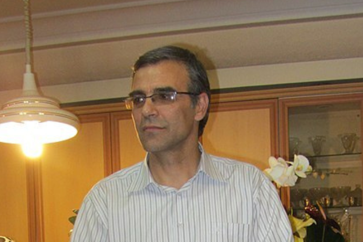 A man with short gray hair and glasses, wearing a light striped shirt, stands indoors near a hanging light and wooden cabinets with glass shelves.