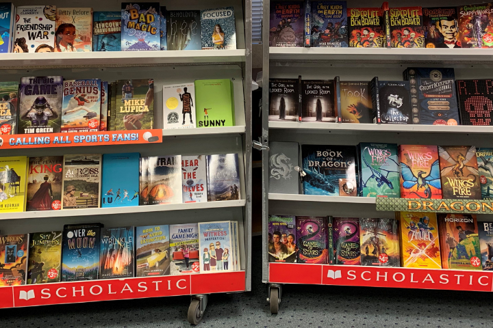 Bookshelves display a variety of colorful children’s and young adult books at a Scholastic book fair, with book covers facing forward and red Scholastic banners along the bottom of each shelf.
