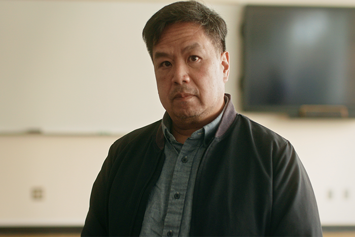 A man with short dark hair wearing a blue shirt and black jacket stands indoors in front of a blurred whiteboard and television screen. He has a neutral expression.