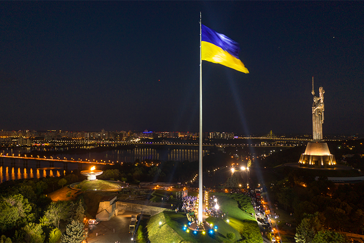 A large Ukrainian flag on a tall pole flies at night in Kyiv, with city lights, a river, and the illuminated Motherland Monument visible in the background.