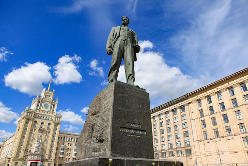 A low-angle view of a large statue of a man standing on a stone pedestal, surrounded by historic buildings, with a bright blue sky and scattered white clouds in the background.