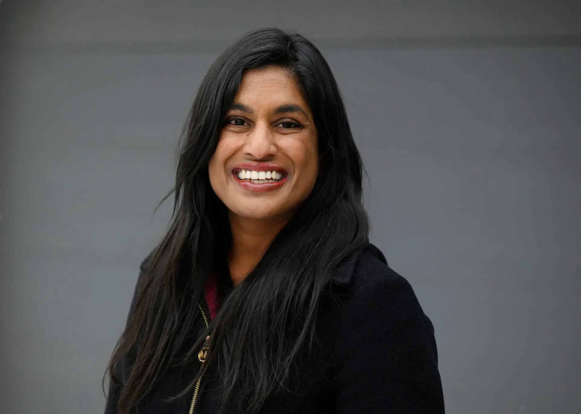 A woman with long black hair and a black coat smiles warmly at the camera, standing in front of a plain gray background—reminiscent of a Vauhini Vara interview portrait.