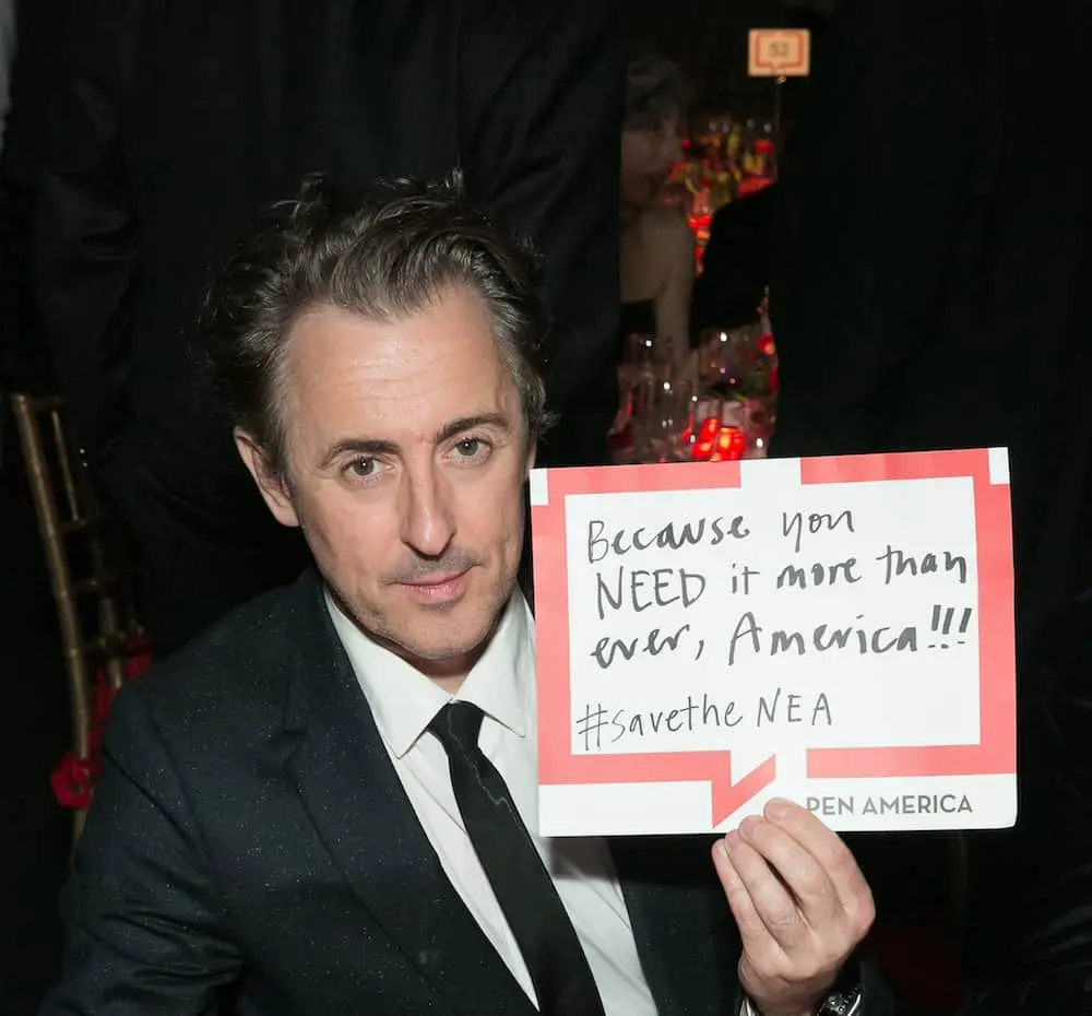 A man in a suit holds a handwritten sign that says, Because you NEED it more than ever, America!!! #savetheNEA at a formal event.