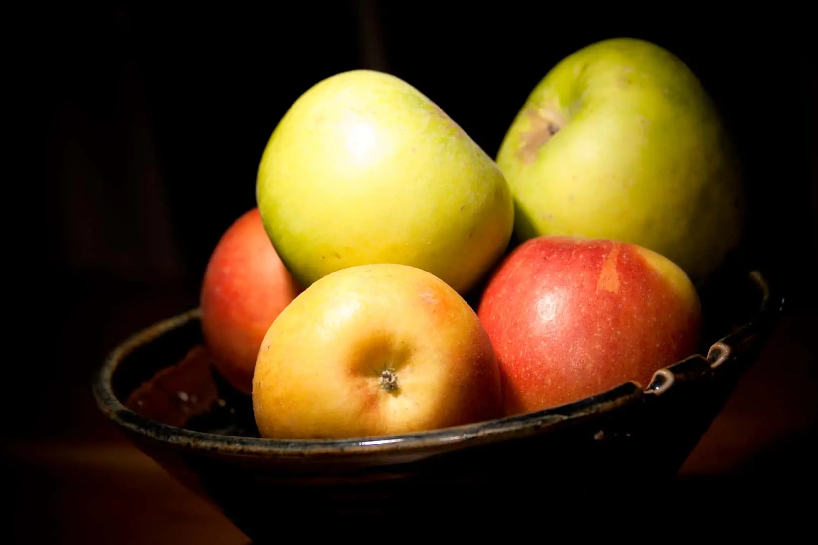 A ceramic bowl filled with a variety of apples, including green and red ones, sits on a dark surface, illuminated by a warm light source that highlights the fruits glossy skin.