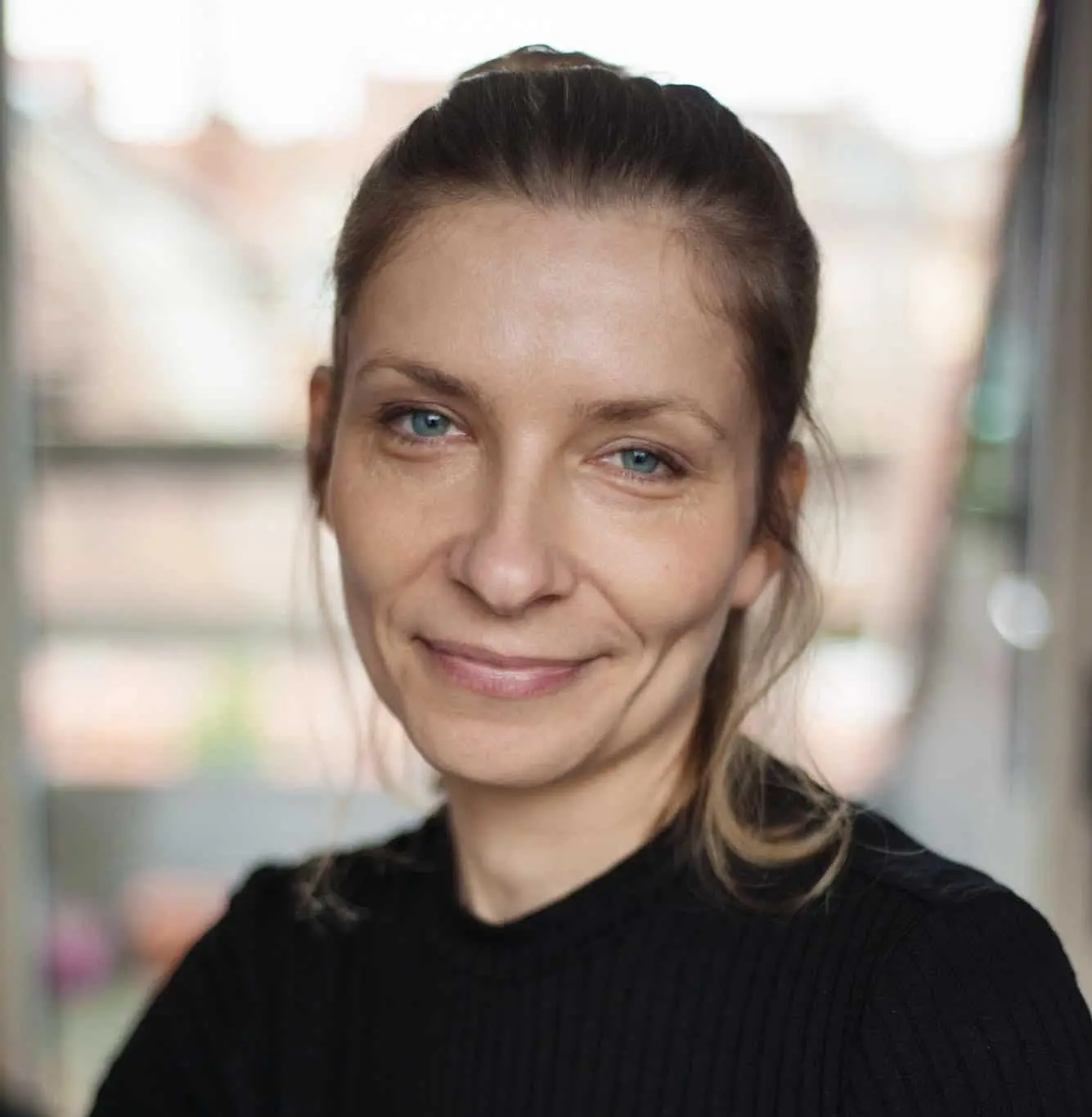 A woman with light hair tied back, wearing a black top, smiles softly at the camera in a softly lit indoor setting with a blurred background, as if ready for a pen ten interview.