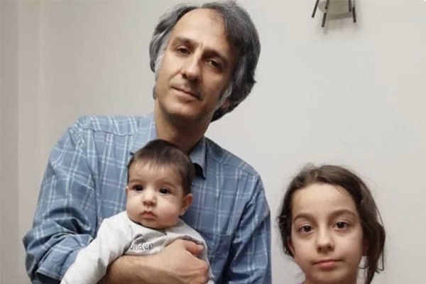 A man in a blue plaid shirt holds a baby in his arms, standing next to a young girl with long brown hair. All three face the camera against a plain indoor background.