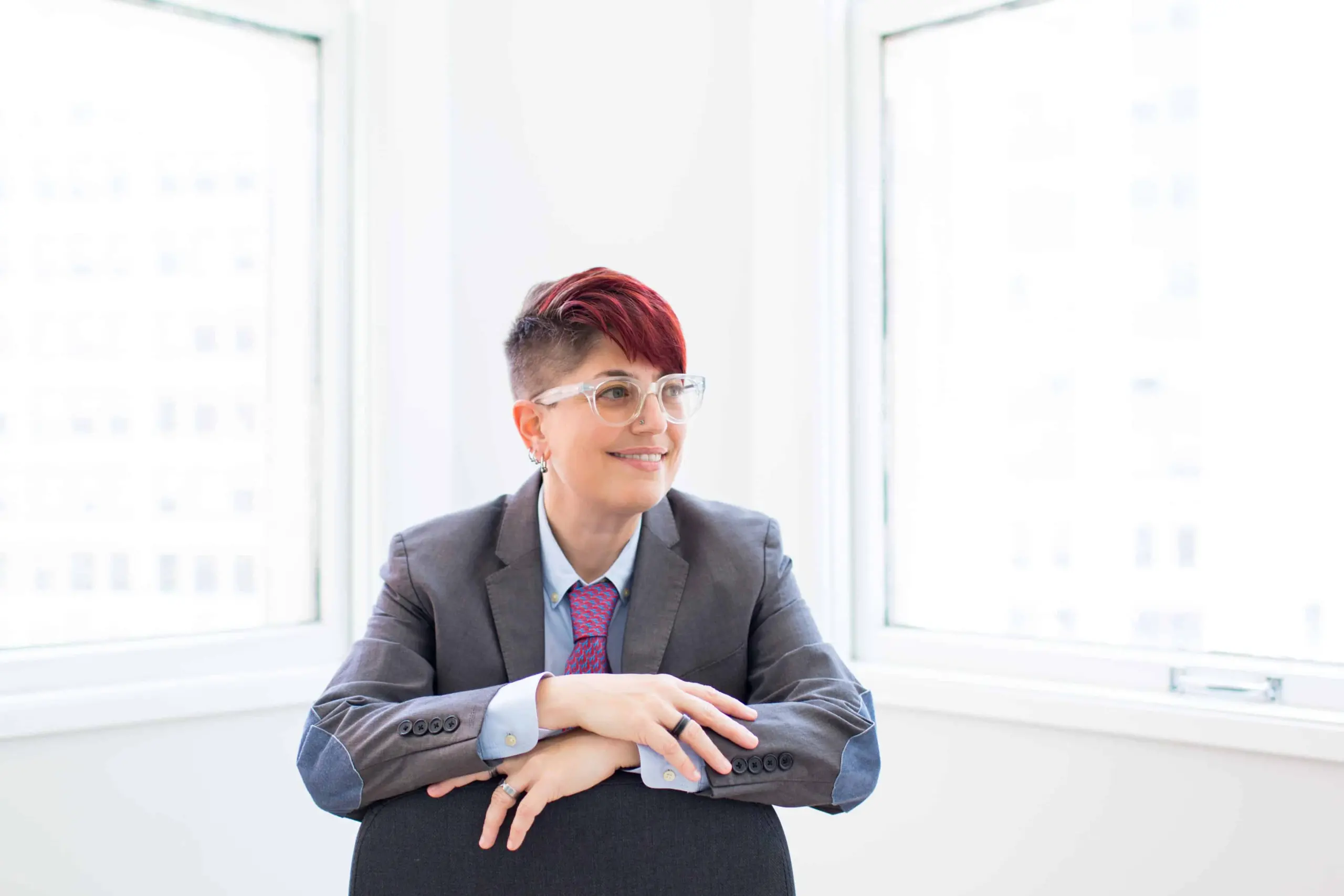 A person with short, red hair and glasses, wearing a gray suit and pink patterned tie, sits smiling with arms resting on the back of a chair in a bright room with two large windows, ready for their pen ten interview.