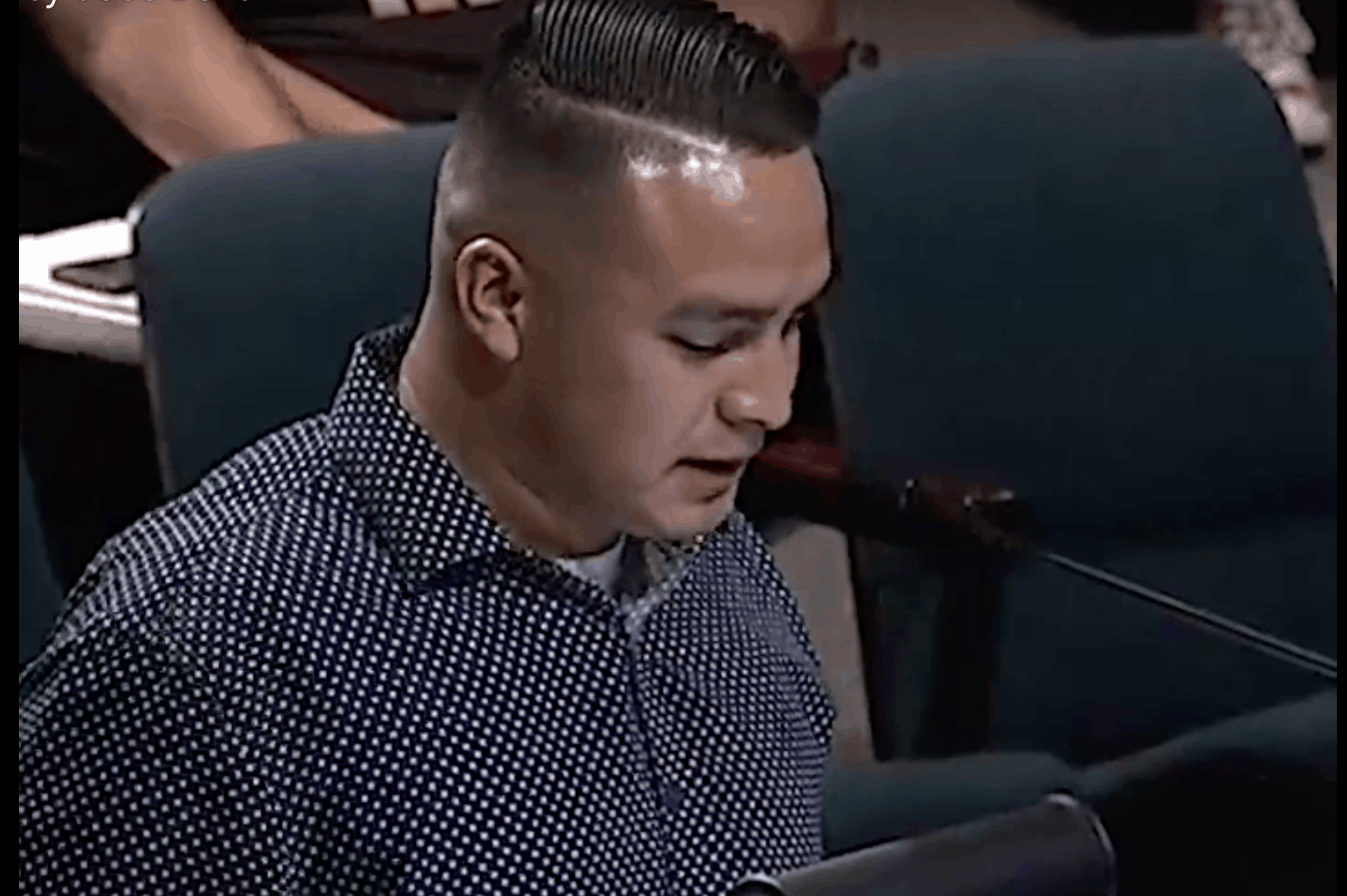 A man with short, slicked-back hair and a dark polka-dot shirt speaks into a microphone at a public meeting, seated in a room with teal chairs.
