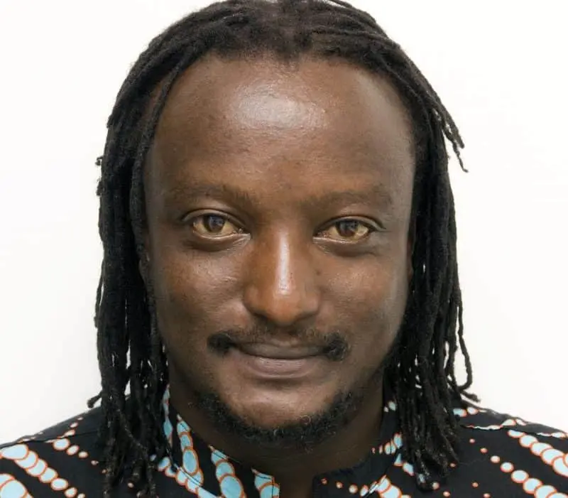A man with medium-length dreadlocks smiles softly at the camera. He is wearing a patterned black shirt with light blue and white dots. The background is plain white.