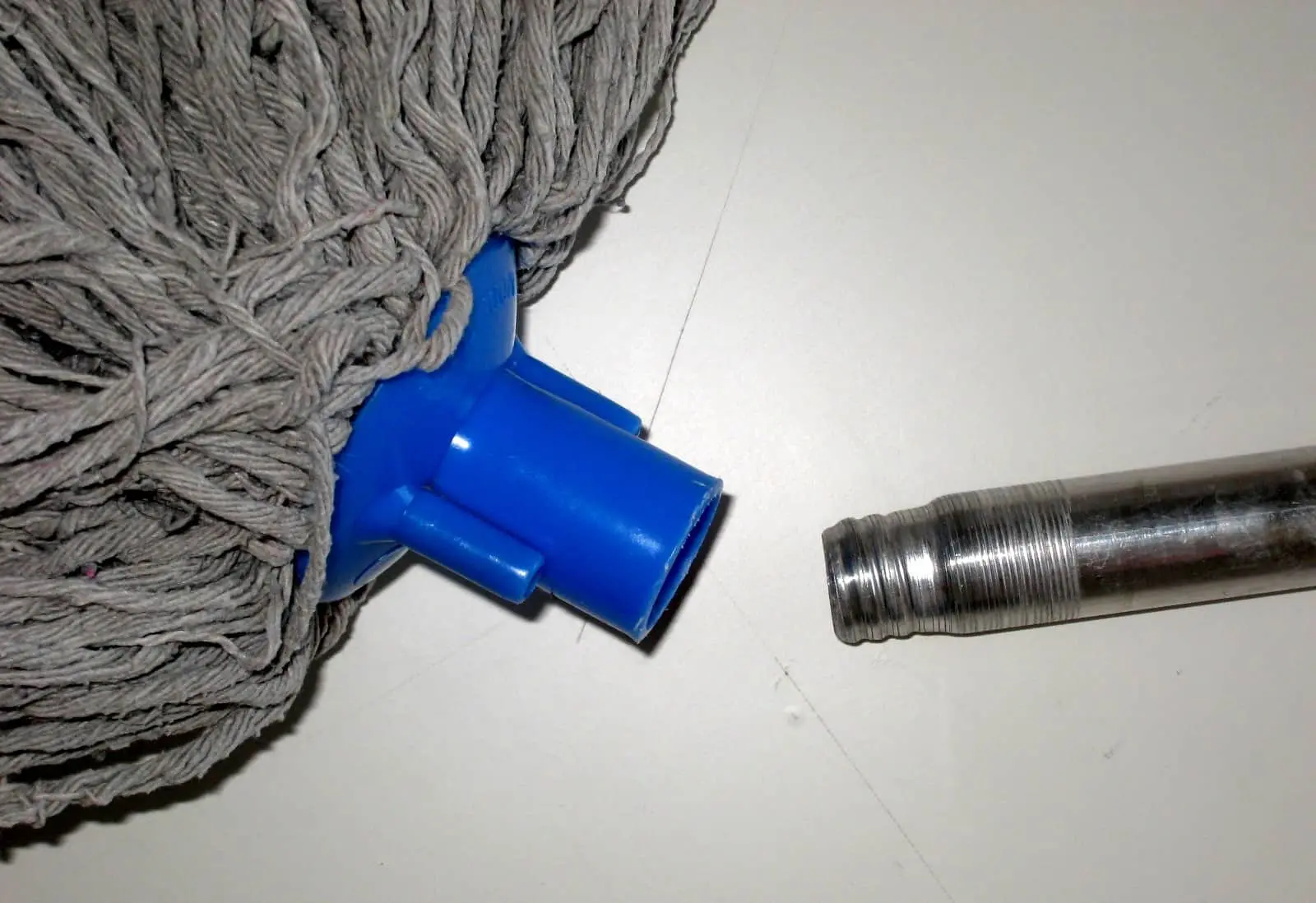 A close-up of a mop head with grey strands and a blue plastic connector next to a detached metal mop handle on a white surface.