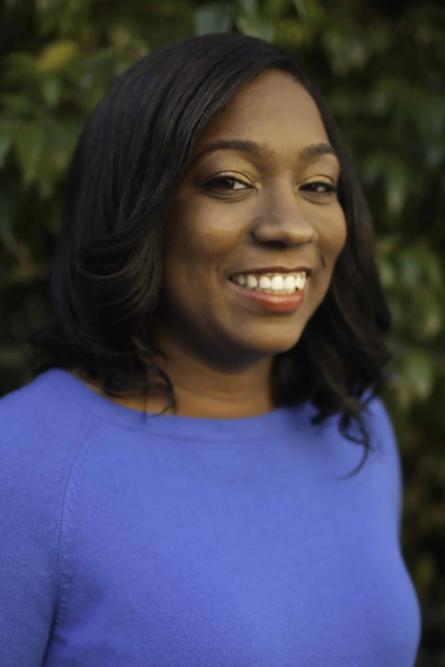 A woman with dark hair wearing a bright blue top smiles warmly at the camera, standing in front of a background of green leaves.