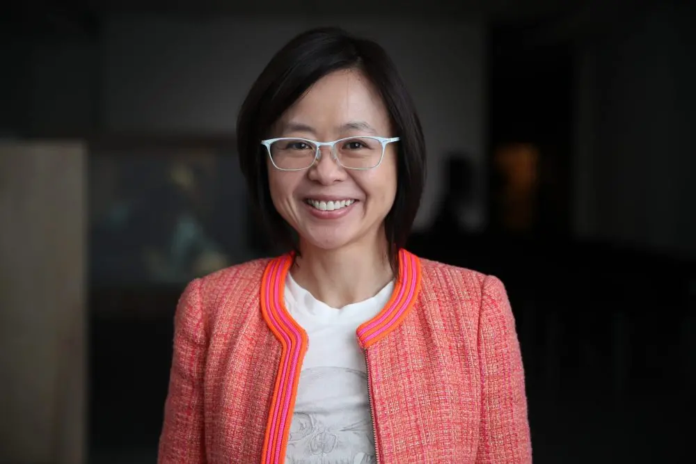 A woman with short dark hair and glasses smiles at the camera. She is wearing a white top and a textured coral jacket with orange trim, standing indoors with a dark, blurred background, holding "tiger heads all the way down by Claire Lin.