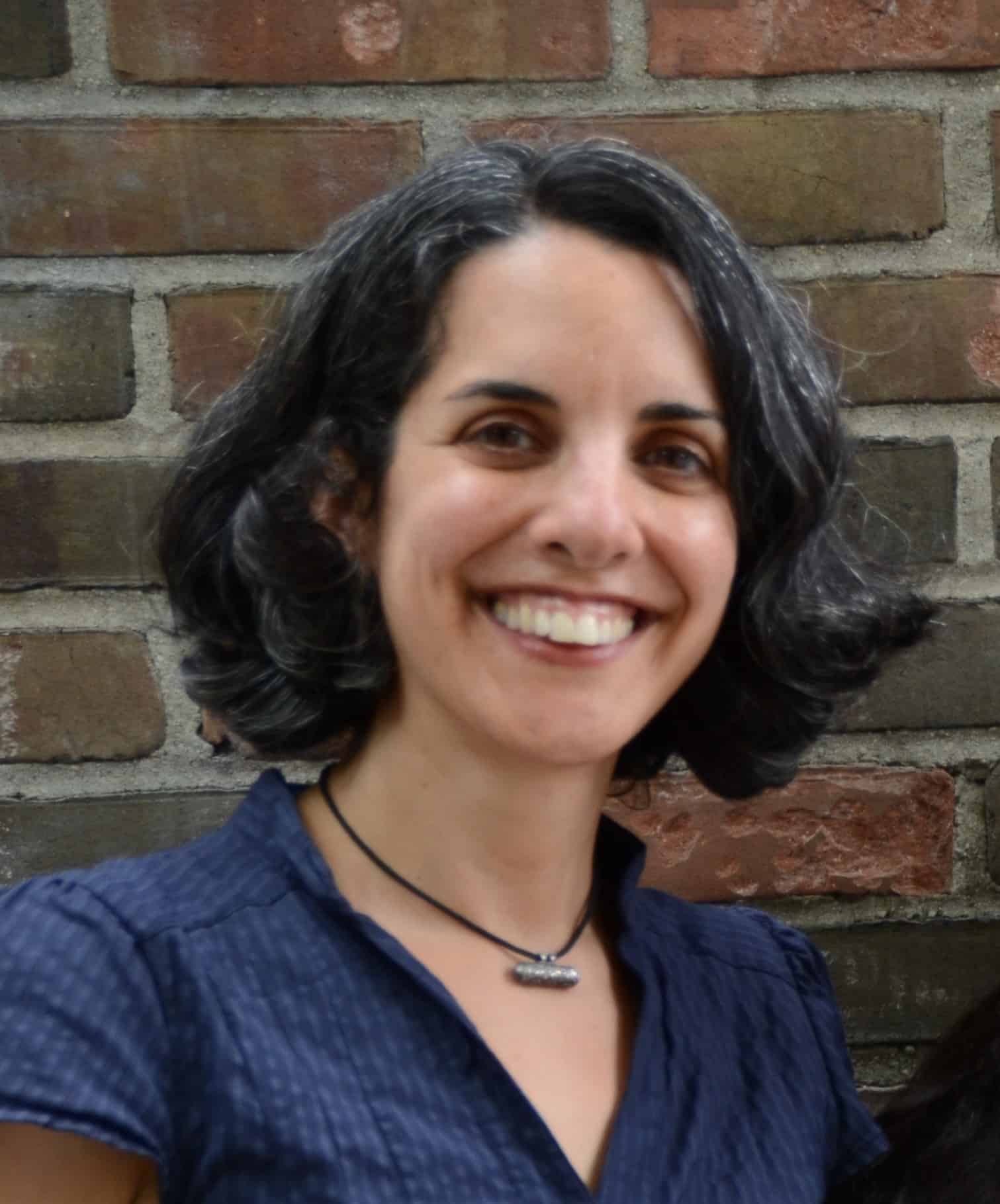A woman with short, curly dark hair with gray streaks smiles in front of a brick wall, reminiscent of Azar Nafisi’s thoughtful presence. She is wearing a dark blue blouse and a black necklace.