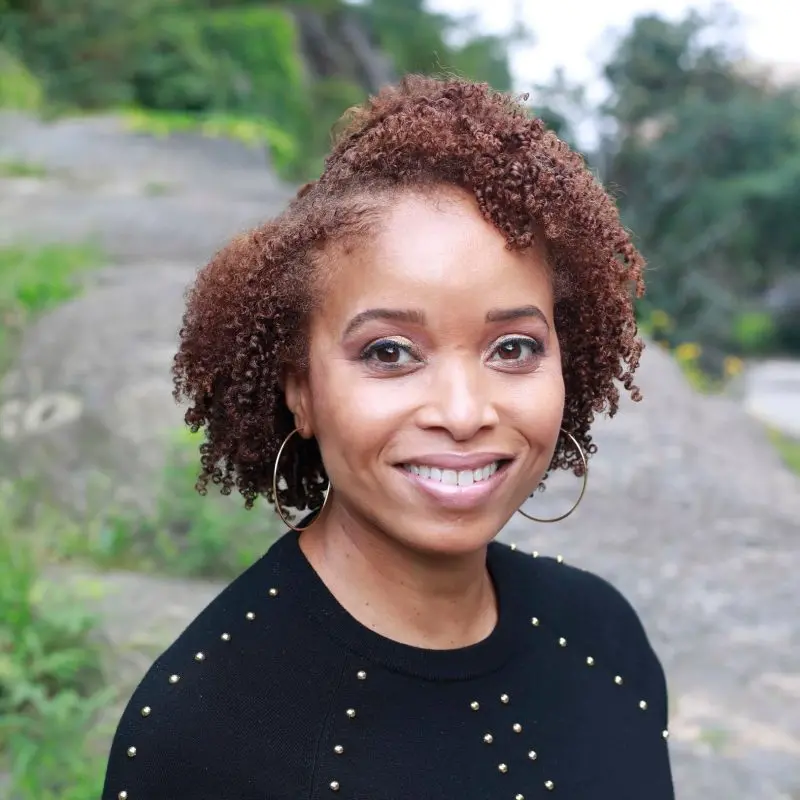 A woman with curly, short brown hair and hoop earrings smiles outdoors, wearing a black sweater with gold studs. Green foliage and rocks are in the background.