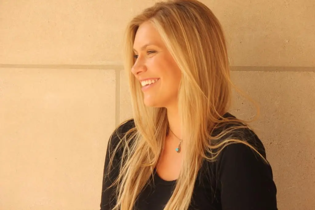 A woman with long blonde hair, wearing a black top and a turquoise pendant necklace, smiles while looking to her left against a light beige wall.