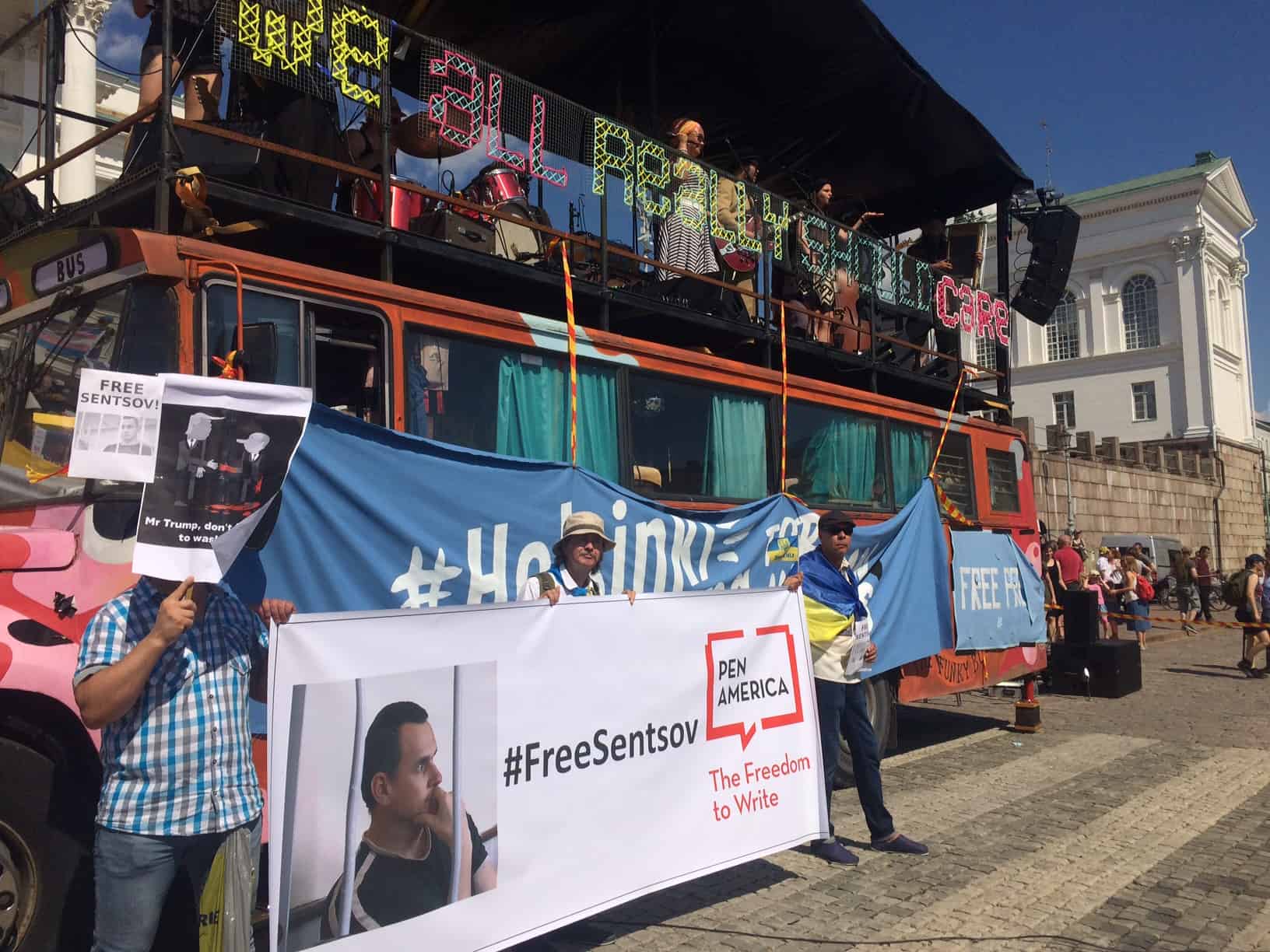 Three people hold a PEN America #FreeSentsov banner in front of a decorated double-decker bus at an outdoor event. One holds a poster with a mans photo; the bus displays colorful signs and a large blue banner.