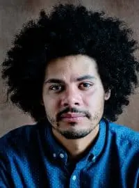 A man with curly black hair and a beard wears a blue button-up shirt and looks directly at the camera against a brown background.