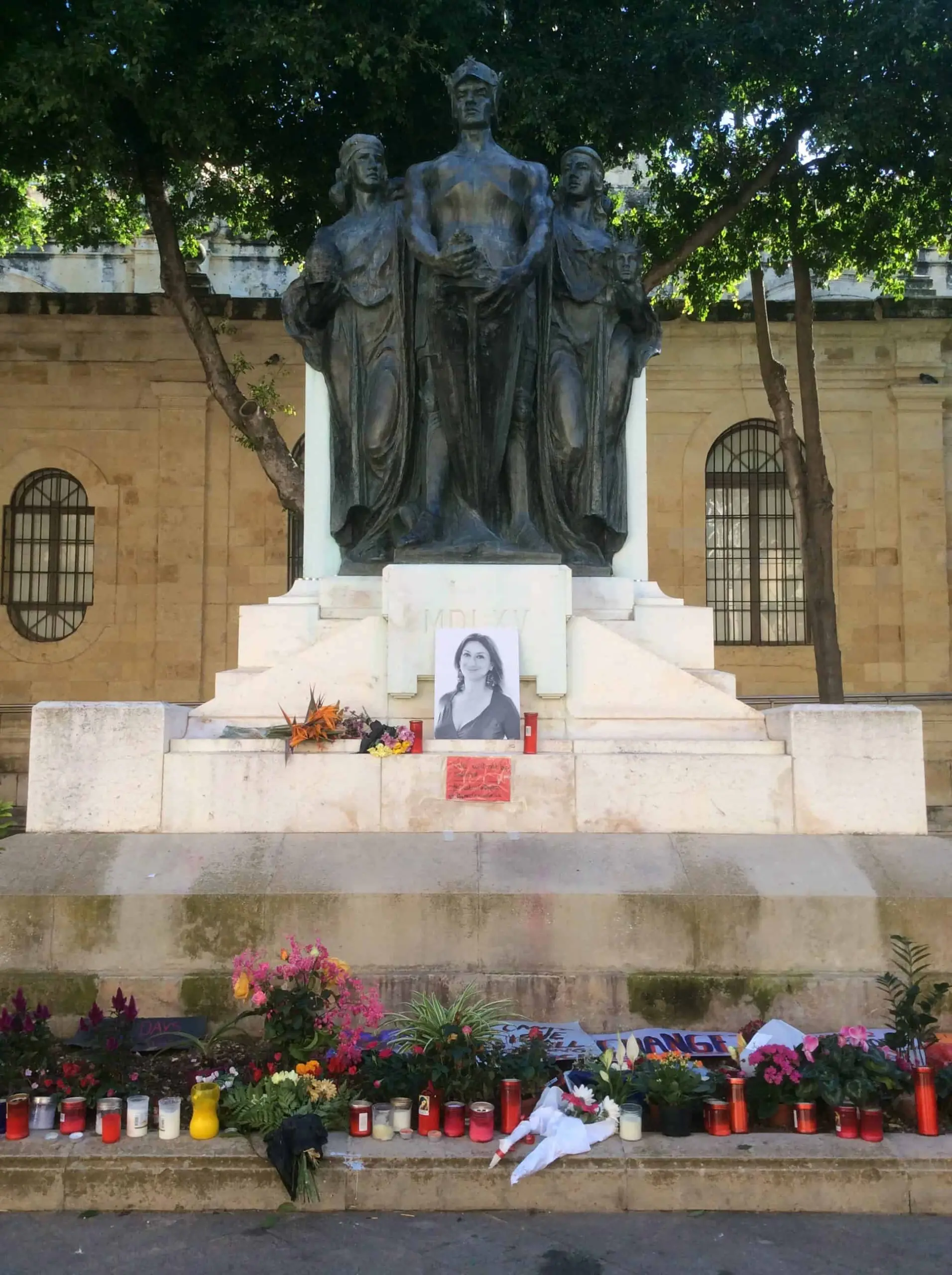 A stone and bronze monument with three standing figures. Flowers, candles, and a black-and-white photo of a woman are placed at its base, suggesting a memorial or tribute. Trees and a building are in the background.