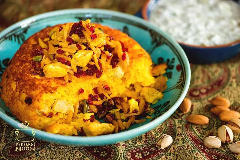 A bowl of Persian Tahchin, a golden baked rice cake with saffron, chicken, barberries, and nuts, sits on a patterned Heim cloth. A side dish of yogurt-cucumber sauce is in the background. Almonds are scattered nearby.