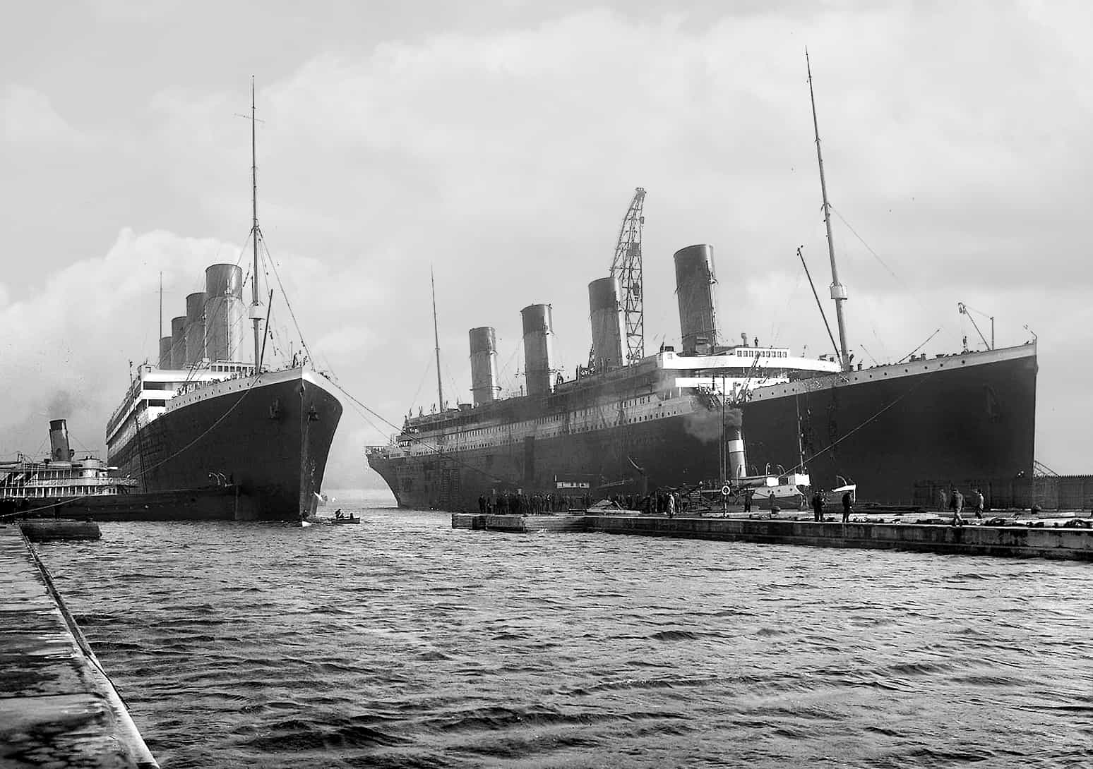 Two large ocean liners docked side by side at a shipyard; workers and cranes are visible on the pier near the ships on a cloudy day.