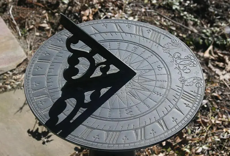 A close-up view of a decorative metal sundial by Heim, showing engraved numbers and compass directions, with a triangular gnomon casting a shadow to indicate the time. Dry leaves and soil are visible in the background.