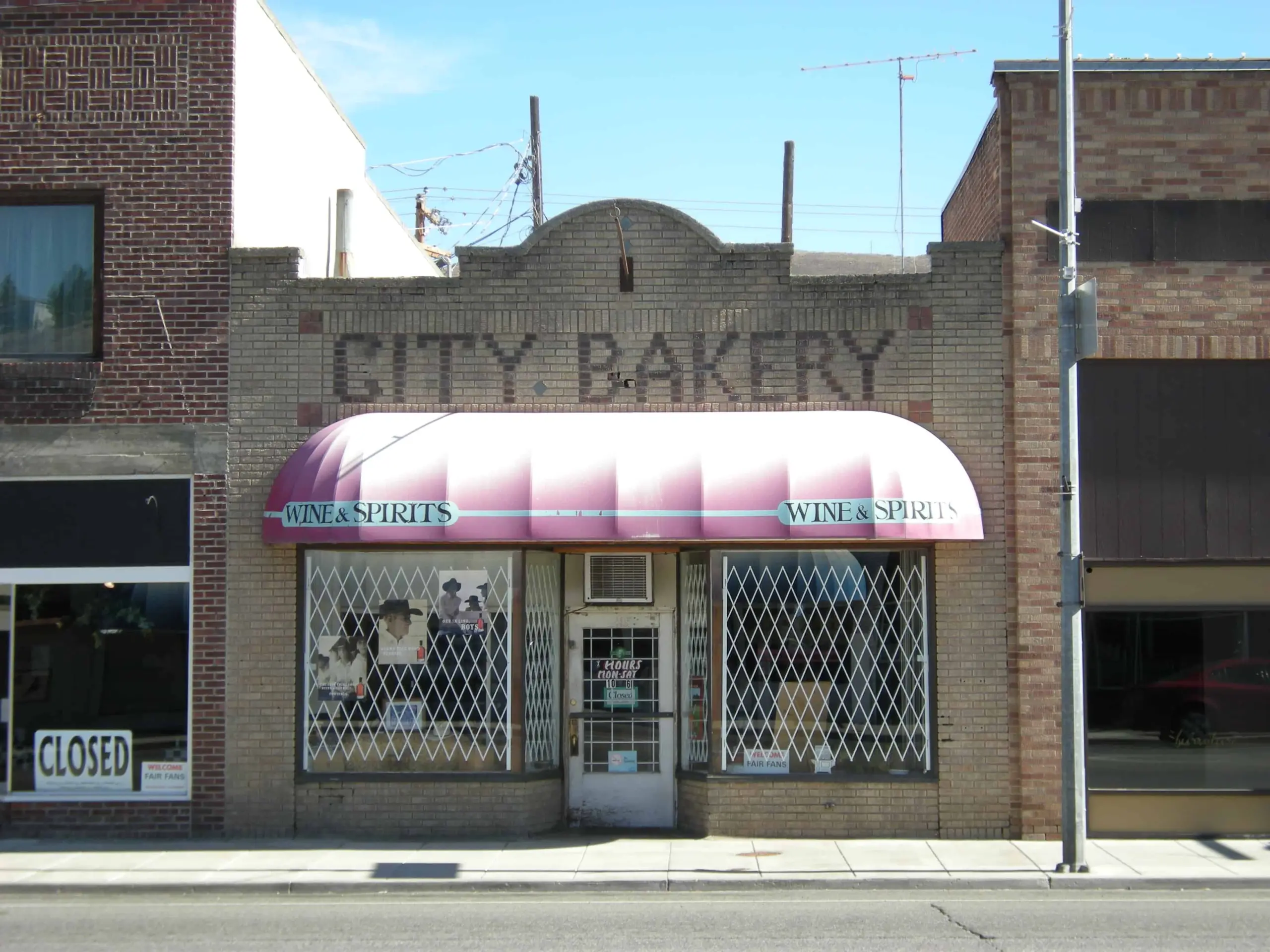 A brick storefront with a faded City Bakery sign above a pink-and-white awning labeled Wine & Spirits. Security bars line the windows and door, hinting at Heim’s cautious charm. A Closed sign hangs in the adjacent shop’s window.