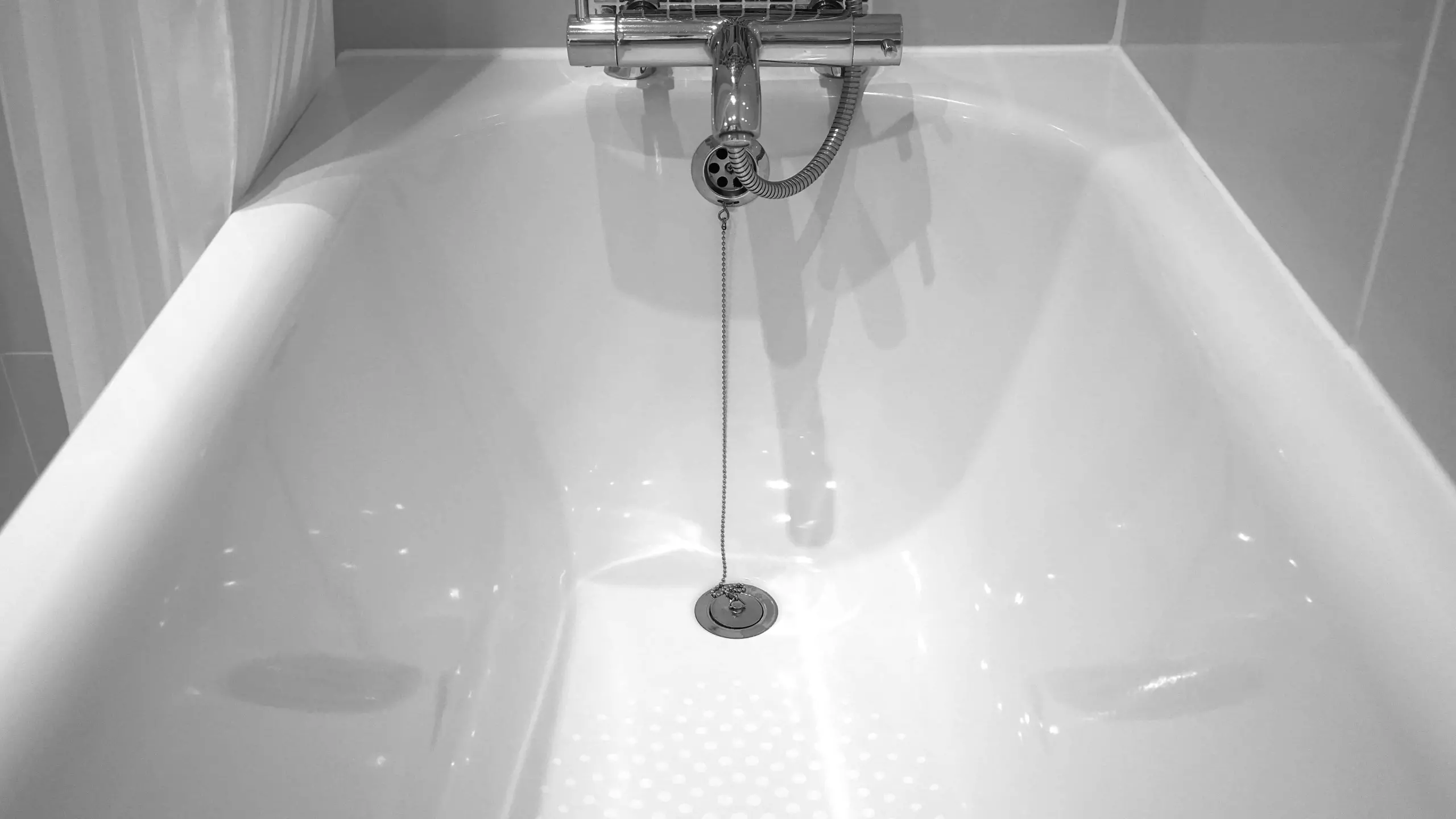 A clean, empty white Heim bathtub with a chrome drain plug attached to a chain and a silver faucet above. The walls around the tub are tiled in light-colored tiles.