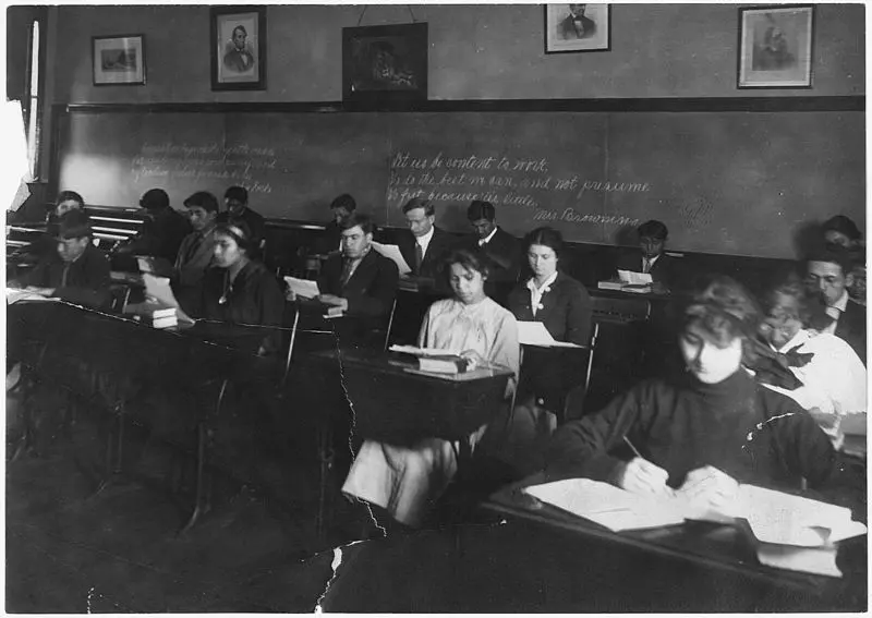 Black and white photo of students in a classroom, sitting at desks and reading or writing. There are chalk writings on the back wall and framed portraits above the blackboard. The classroom appears to be from the early 20th century.
