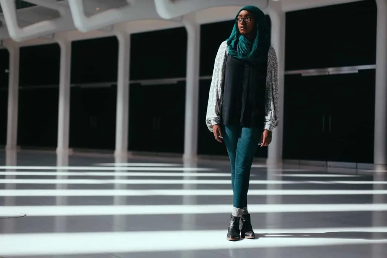 A woman wearing a teal headscarf, glasses, patterned sweater, and jeans stands in a spacious, modern building with bright sunlight casting striped shadows across the floor.