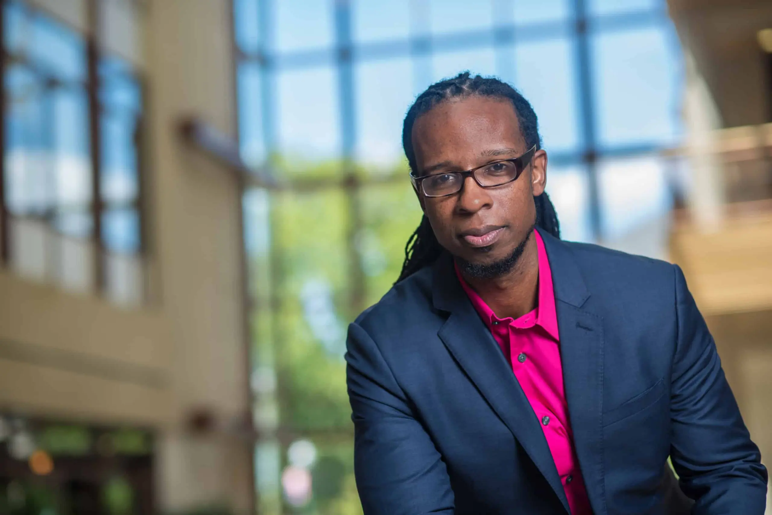 A man with glasses and long braids, wearing a navy blazer over a bright pink shirt, stands indoors in front of large windows with natural light streaming in.