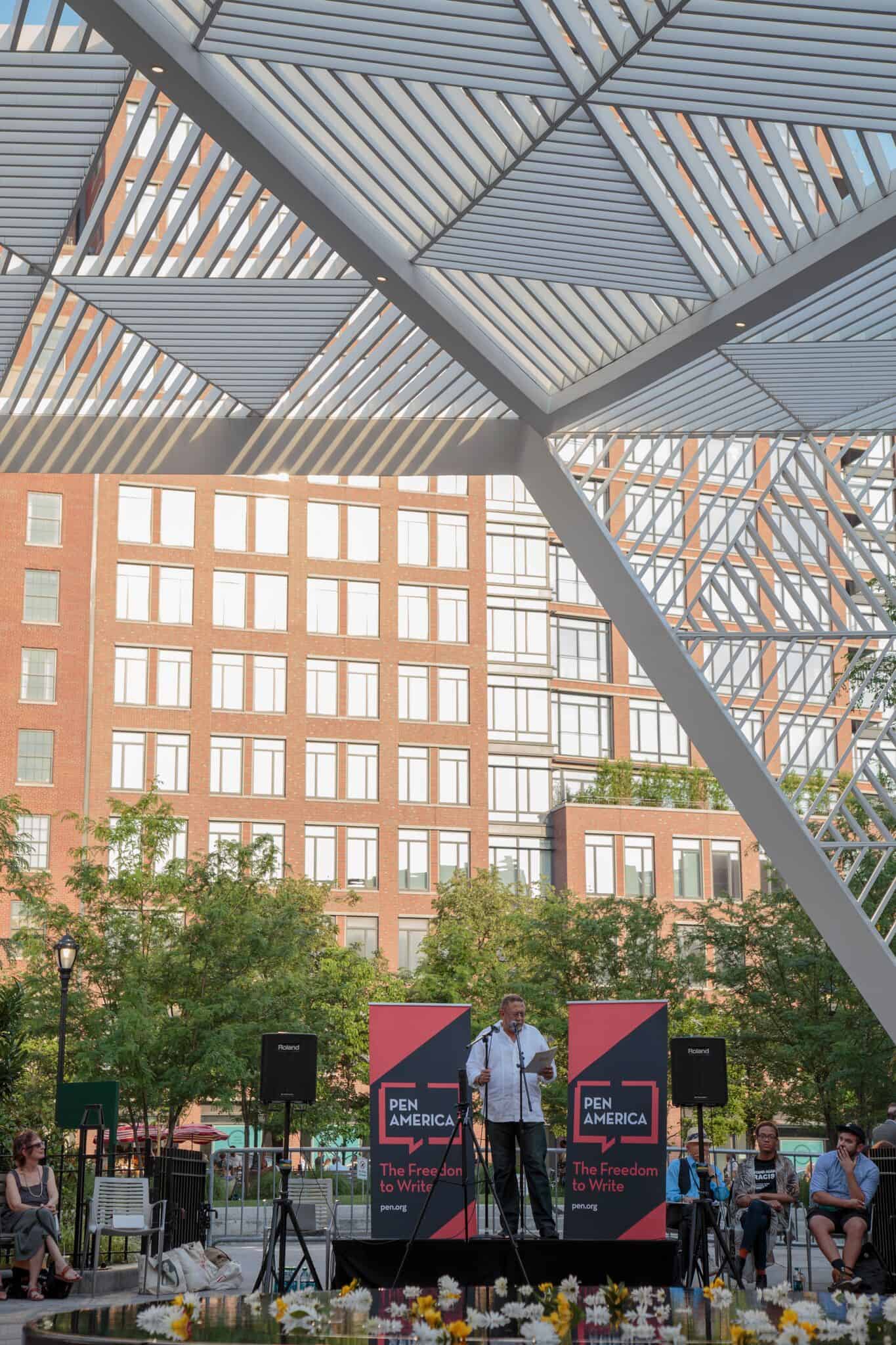 A speaker stands at a podium with two PEN America: The Freedom to Write banners, addressing an outdoor audience under a modern pavilion, with a city building and trees in the background.