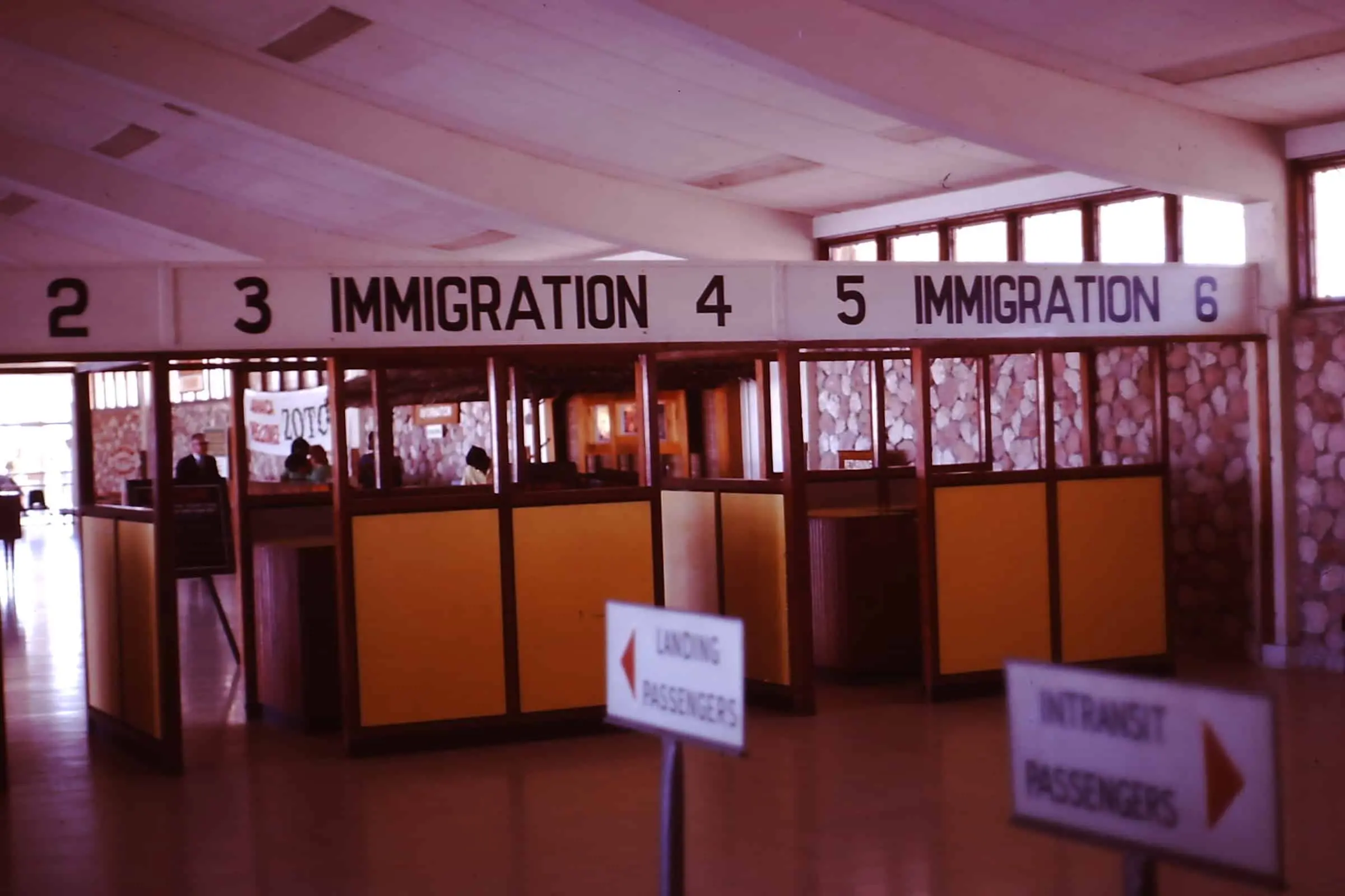 An indoor immigration checkpoint area with numbered booths 2–6, overhead signs marked IMMIGRATION, and directional signs in the foreground for landing and intransit passengers.