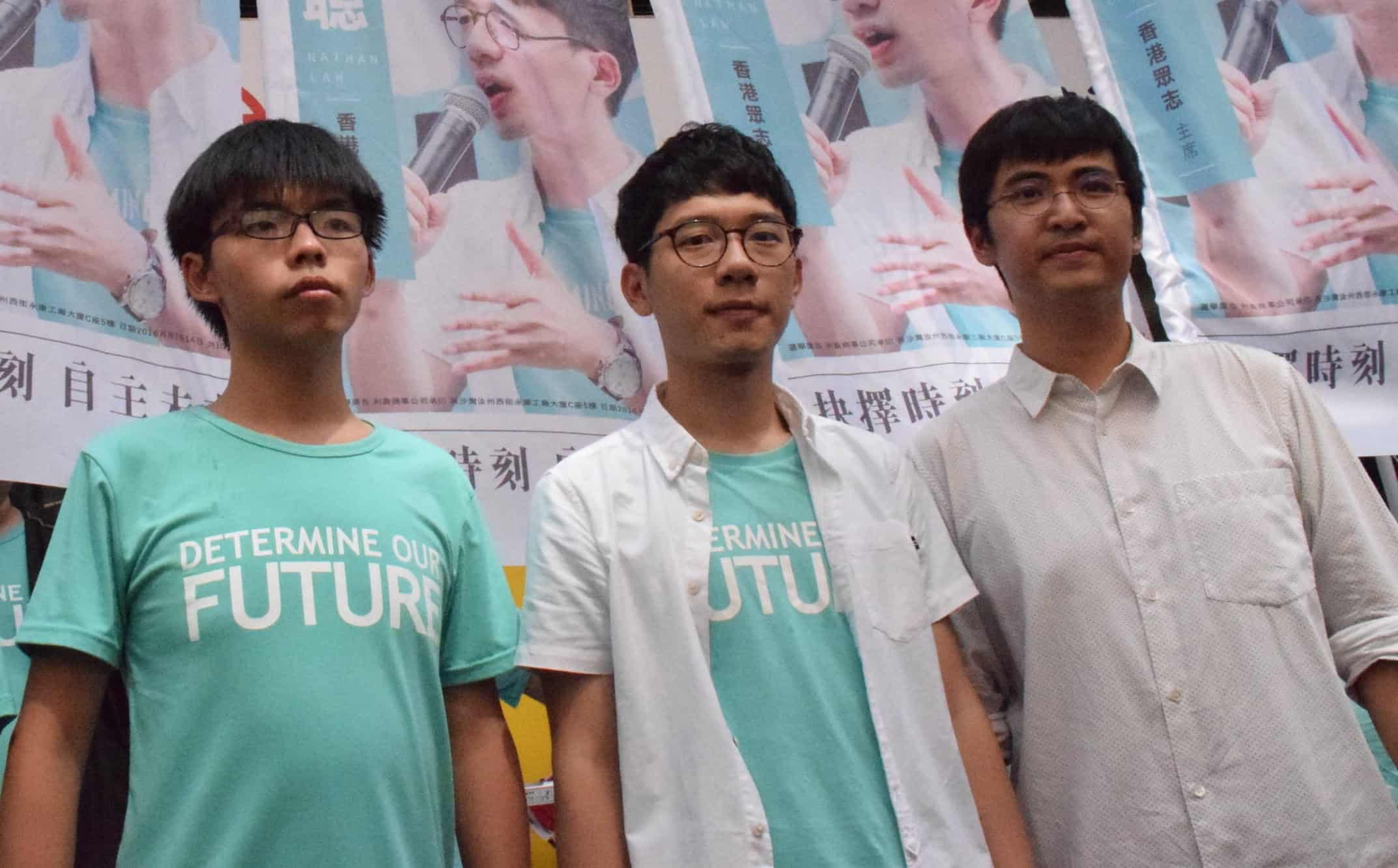 Three young men stand in front of campaign posters. Two wear teal shirts reading DETERMINE OUR FUTURE, while the third wears a white shirt. The background posters feature one of the men speaking.