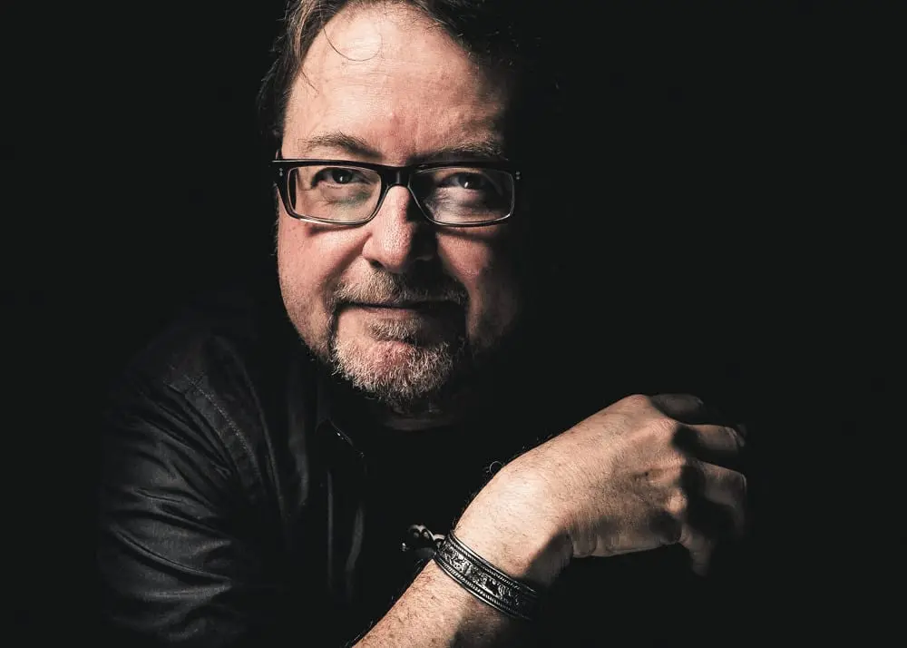 A man with glasses, a short beard, and mustache sits in a dimly lit setting, wearing a dark shirt and a bracelet, with his arm resting across his chest and a slight smile on his face.