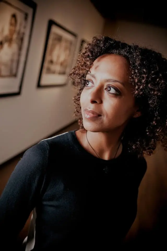 A woman with curly hair and a black top looks thoughtfully to the side while standing in an art gallery with framed pictures on the wall behind her.