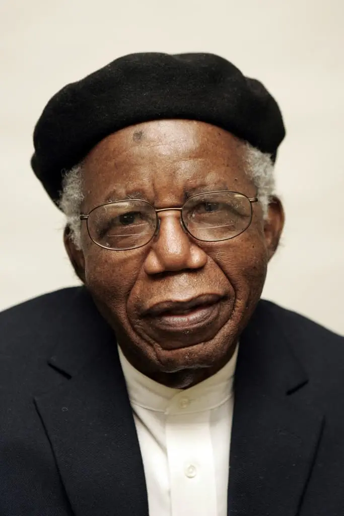 An elderly Black man with gray hair, wearing glasses, a black beret, a dark blazer, and a white shirt, looks directly at the camera against a plain light background.