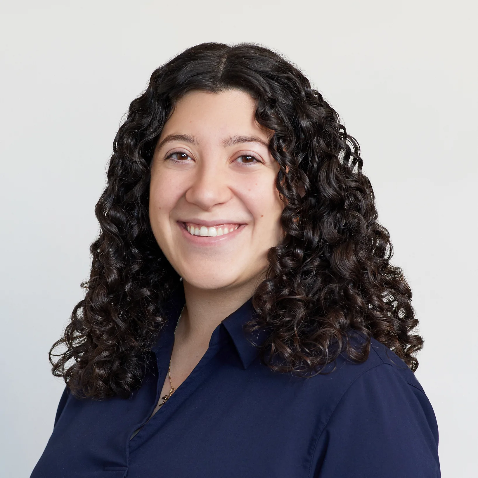 A woman with long, dark, curly hair smiles at the camera. She is wearing a dark navy blouse and is posed in front of a plain, light-colored background.